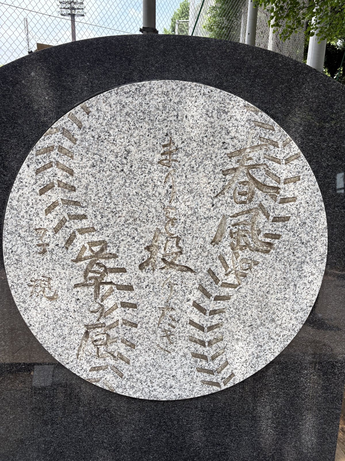 句碑のボール。白い円盤に縫い目が走り、その間に「春風や まりを投げたき 草の原」の句が刻まれている。