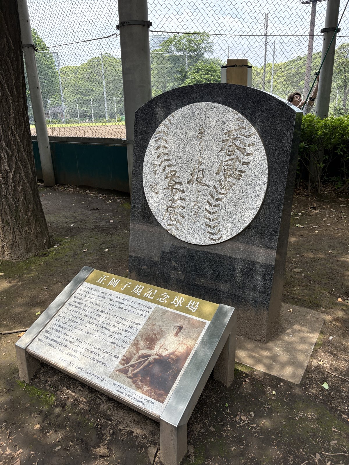 黒い御影石の句碑と、手前の解説板。球場脇の木陰に、ひっそりと立っていた。